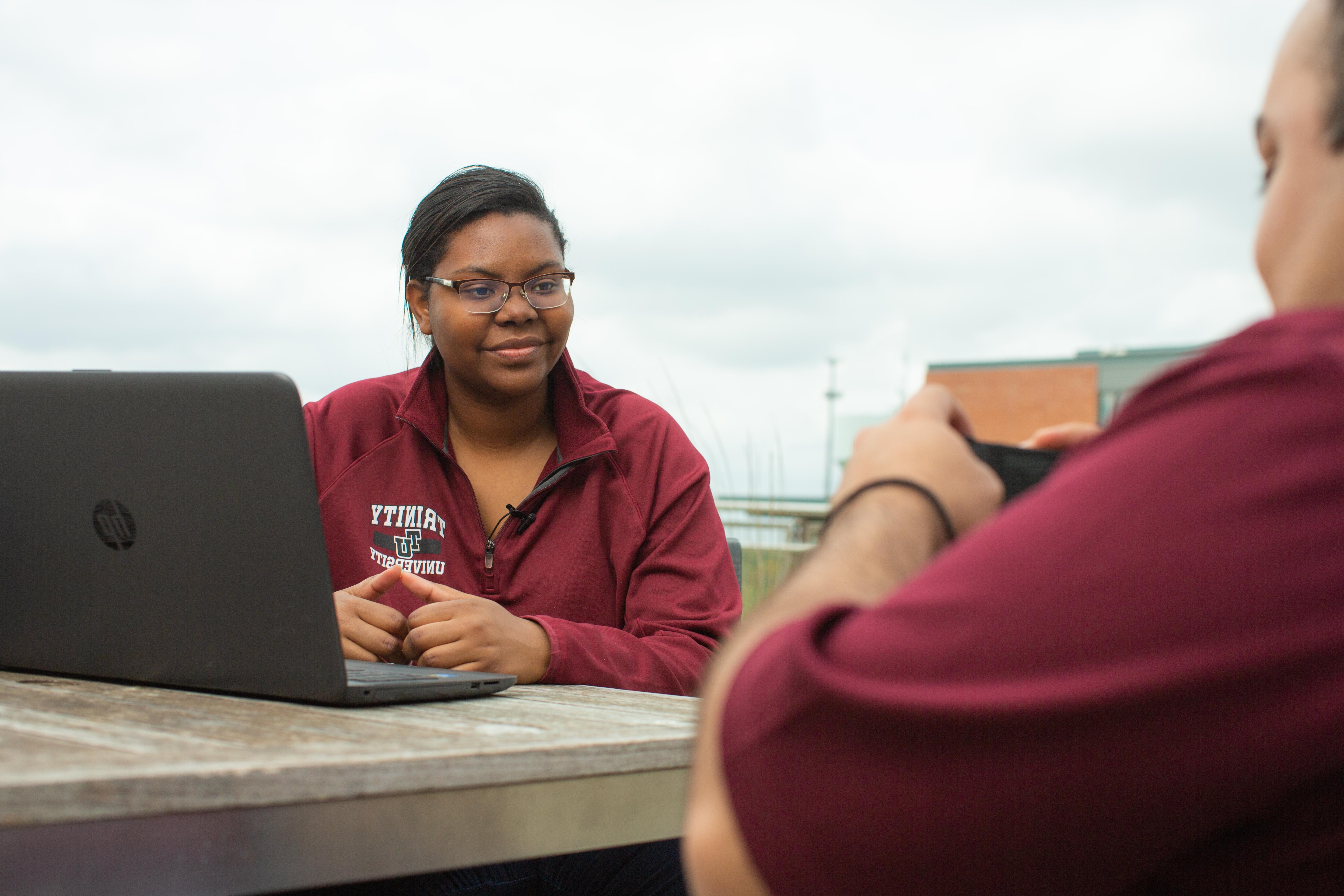 photo of students sitting down, talking and working on a laptop outside